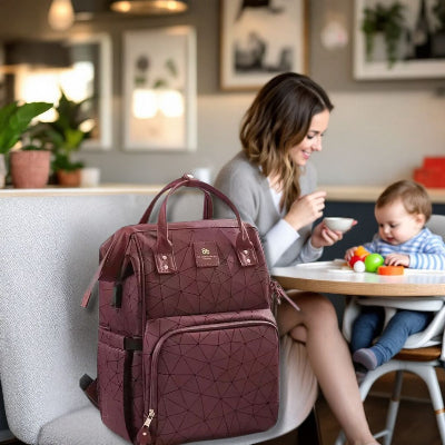 Une maman partageant un moment avec son enfant dans un café chaleureux, avec le sac à dos à langer Bordeaux Chic en arrière-plan, montrant son élégance et son côté pratique.