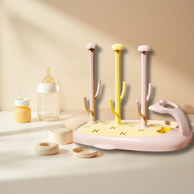 Égouttoir biberon CompactBébé™ aux tons jaune pastel et rose poudré, avec accessoires de bébé autour, parfait pour une utilisation quotidienne avec un séchage rapide et sans humidité résiduelle.