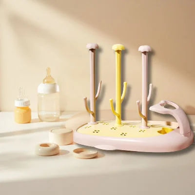 Égouttoir biberon CompactBébé™ aux tons jaune pastel et rose poudré, avec accessoires de bébé autour, parfait pour une utilisation quotidienne avec un séchage rapide et sans humidité résiduelle.