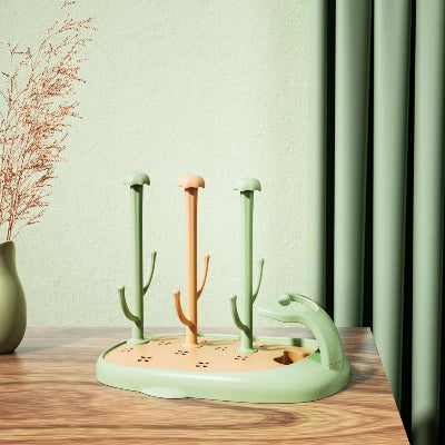 Sèche biberon CompactBébé™ coloris vert menthe et abricot, installé sur une table en bois devant un rideau vert assorti, parfait pour organiser les biberons et tétines dans une cuisine au style naturel et moderne.