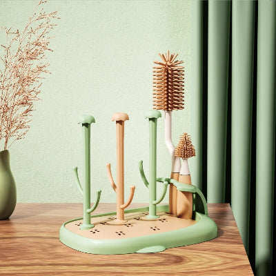 Sèche biberon vert menthe CleanDrop™ avec brosses en silicone positionnées dans leur support latéral, posé sur une table en bois contre un rideau vert pastel.

