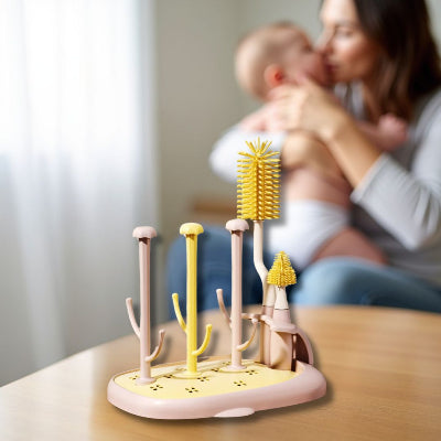 Sèche biberon rose et jaune CleanDrop™ sur table, avec une maman tenant son bébé en arrière-plan, illustrant l’organisation optimale de la biberonnerie à la maison.

