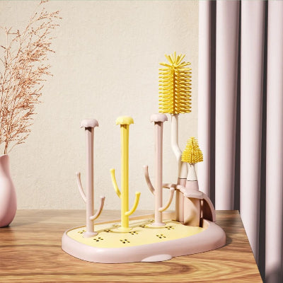 Sèche biberon CleanDrop™ rose pastel et jaune, équipé de brosses à biberon assorties, installé sur une table dans une ambiance douce et lumineuse.

