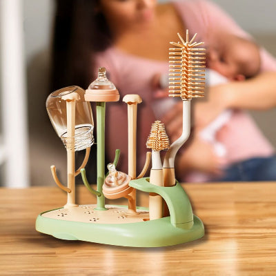 Sèche biberon CleanDrop™ en situation réelle, avec biberons, tétines et brosses disposés, en arrière-plan une maman allaitant son bébé dans une ambiance chaleureuse.

