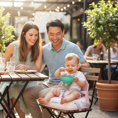 Bébé installé dans un siège repas bébé pliable rose au restaurant, réhausseur de table enfant sécurisé pour partager le repas avec ses parents.
