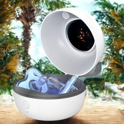 Stérilisateur tétine UV portable ouvert avec sucette à l’intérieur, posé sur une table en bois en extérieur avec palmiers en arrière-plan. Idéal pour stériliser les tétines pendant les sorties en vacances ou à la plage.