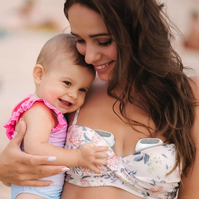 Maman souriante portant un tire-lait mains libres tout en tenant son bébé sur la plage, illustrant une extraction discrète et pratique en extérieur.